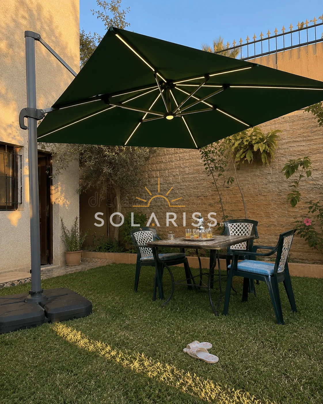 باراسول سولاريس 3×3 متر بالأخضر الملكي وإضاءة LED فوق طاولة شاي في حديقة منزل عند الغروب